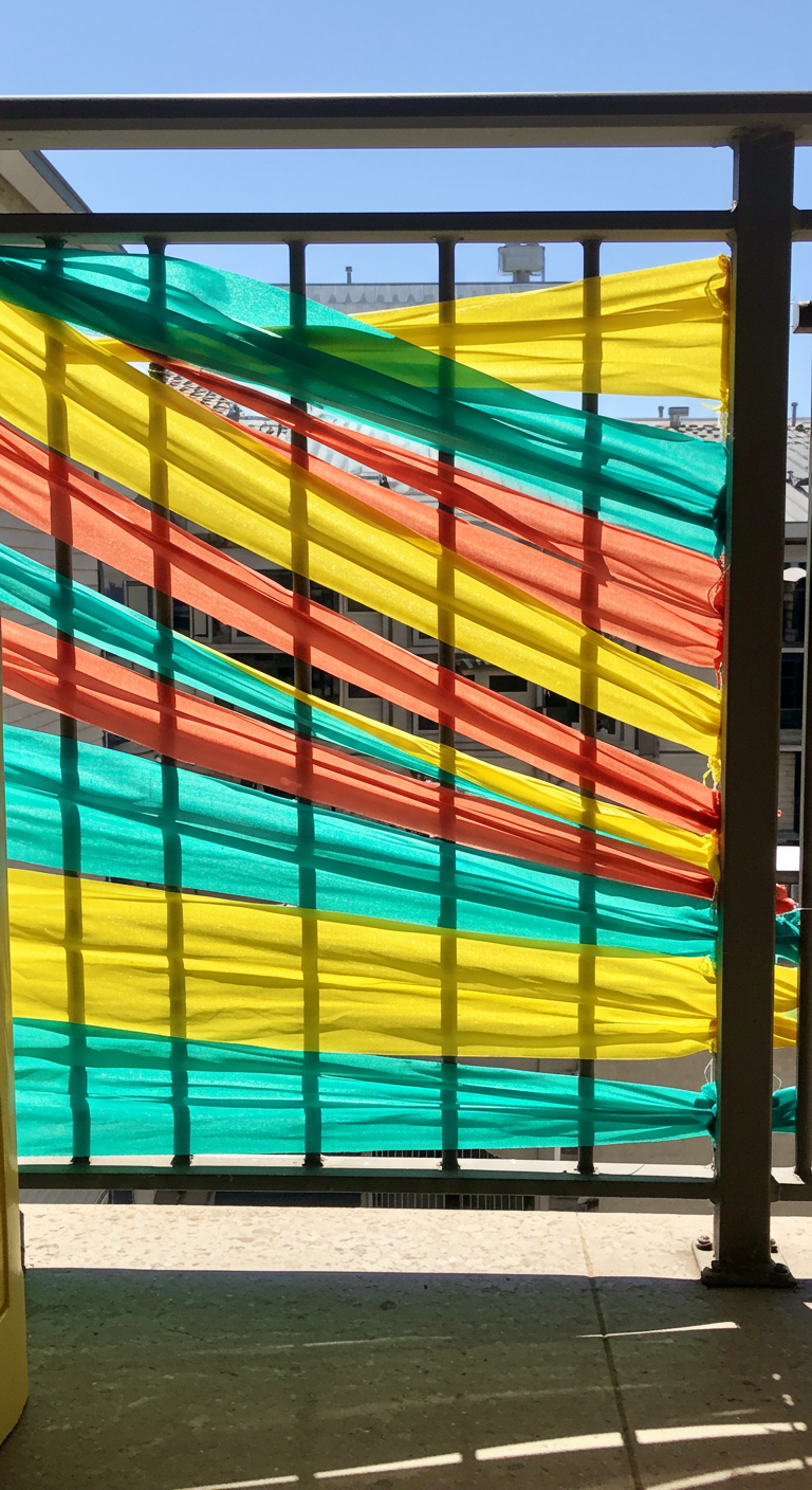 Bunte Stoffbahnen (grün, gelb, orange) sind durch ein Metallgeländer geflochten.