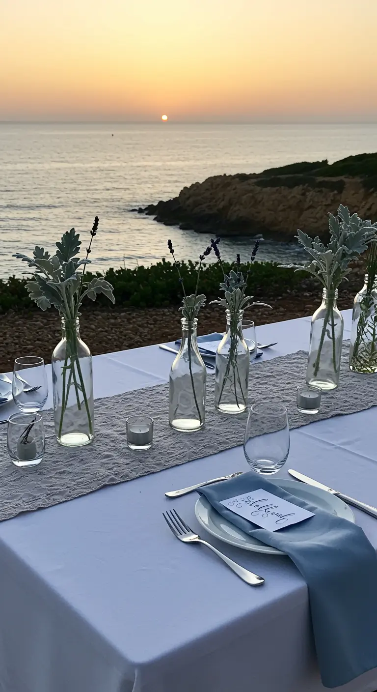 Weiß gedeckter Tisch am Meer mit blauen Servietten und silbergrünen Blättern in Glasflaschen.