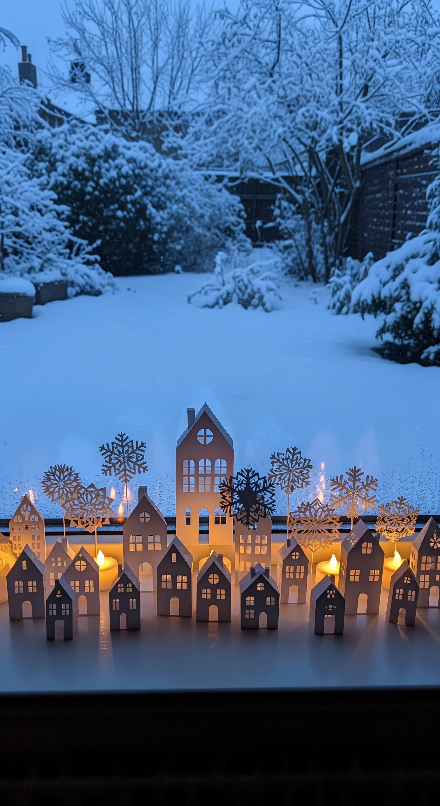Fensterbank dekoriert mit weißen Papierhäusern und Schneeflocken, abendlich beleuchtet.