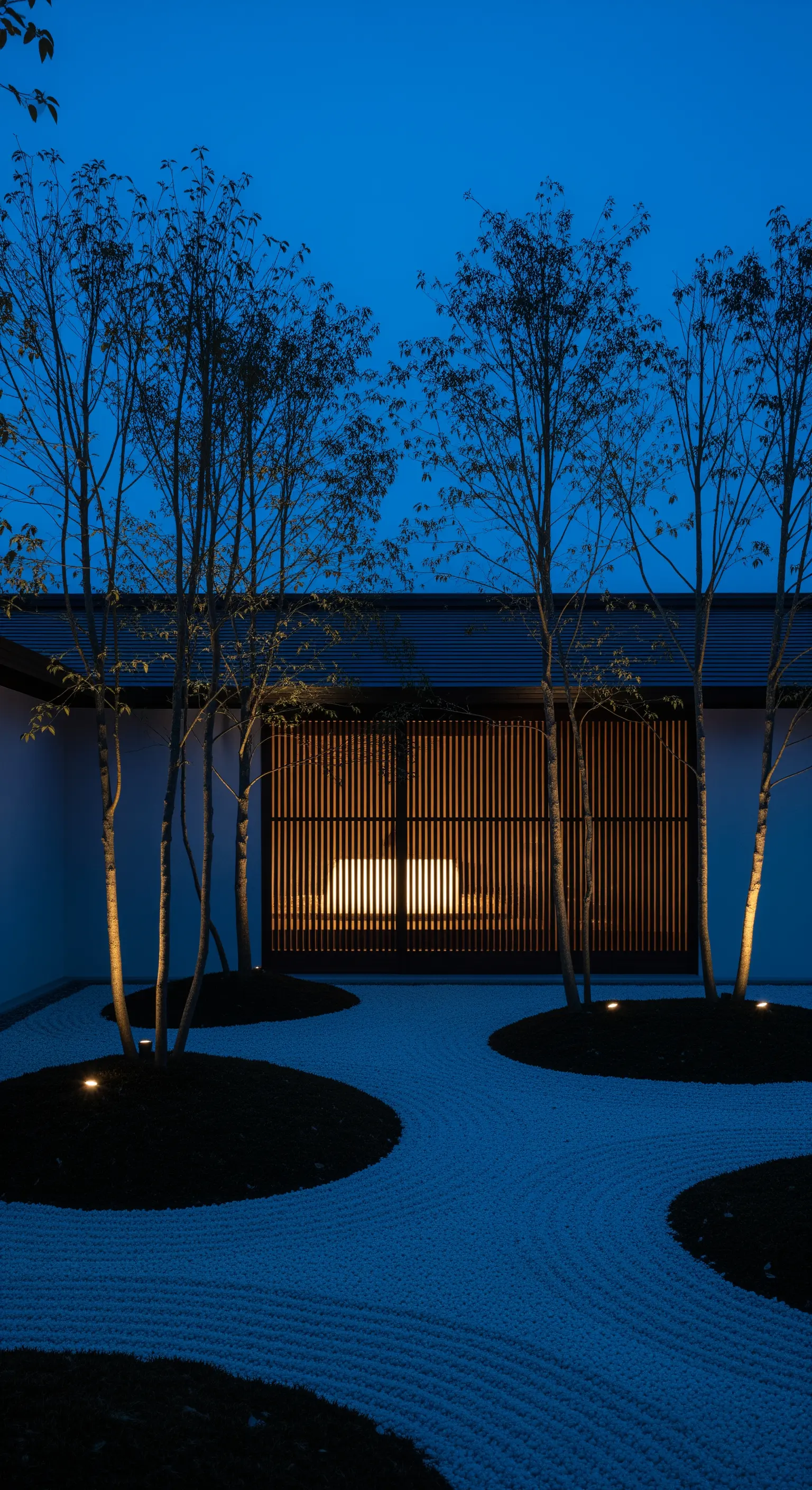 Abendlicher Japandi Hofgarten mit Bäumen, Kies und beleuchtetem Sichtschutz