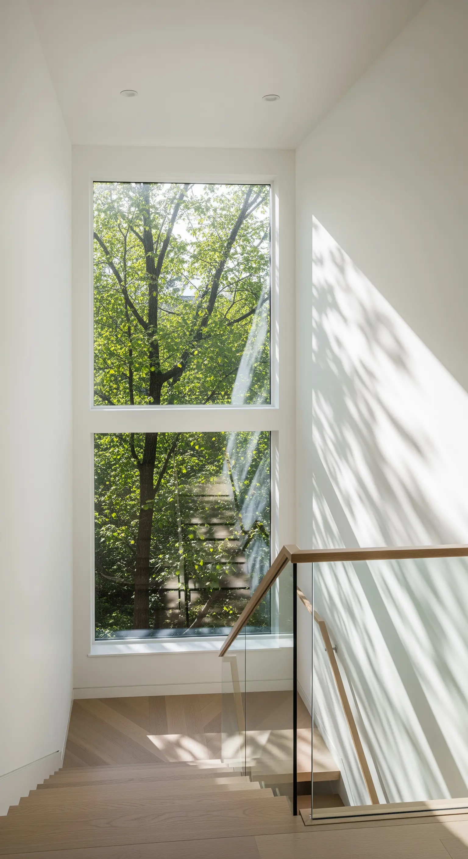 Modernes Treppenhaus aus Holz mit einem hohen, vertikalen Fenster und Blick auf grüne Bäume.