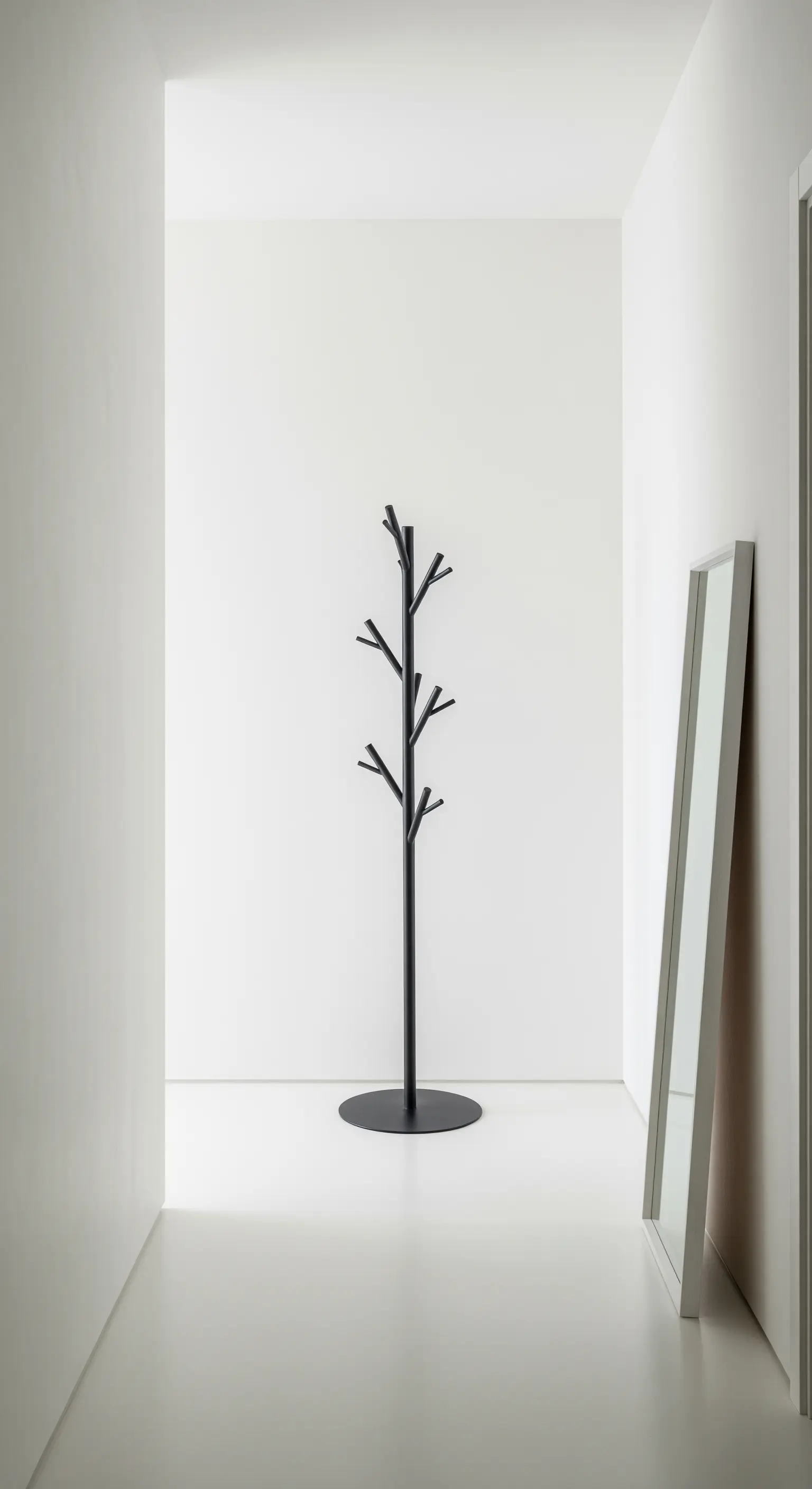 Minimalistischer, baumförmiger Garderobenständer und ein an die Wand gelehnter Spiegel in einem weißen Flur.