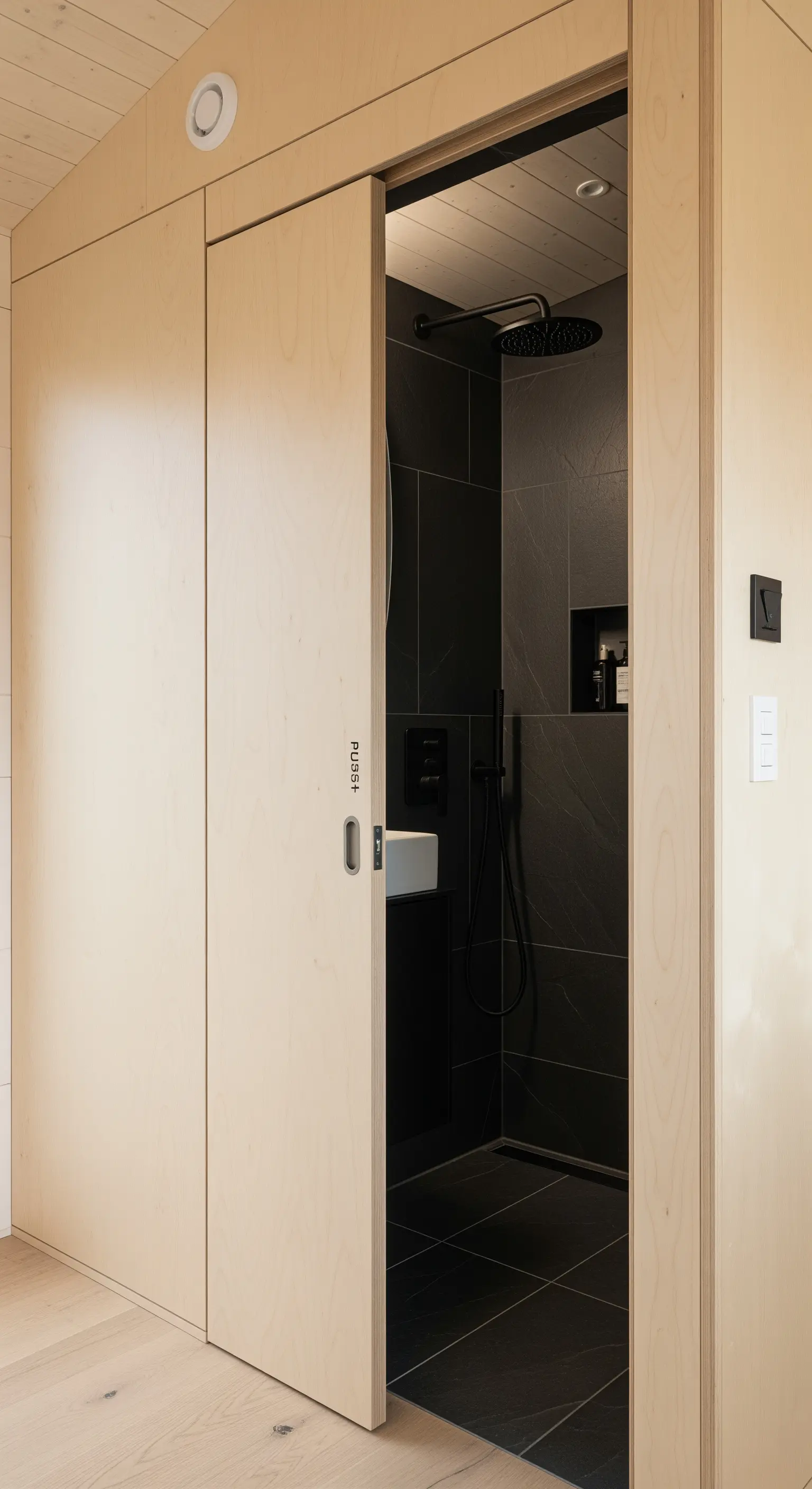 Schiebetür aus hellem Holz, die sich in die Wand integriert und zu einem dunklen Bad führt.