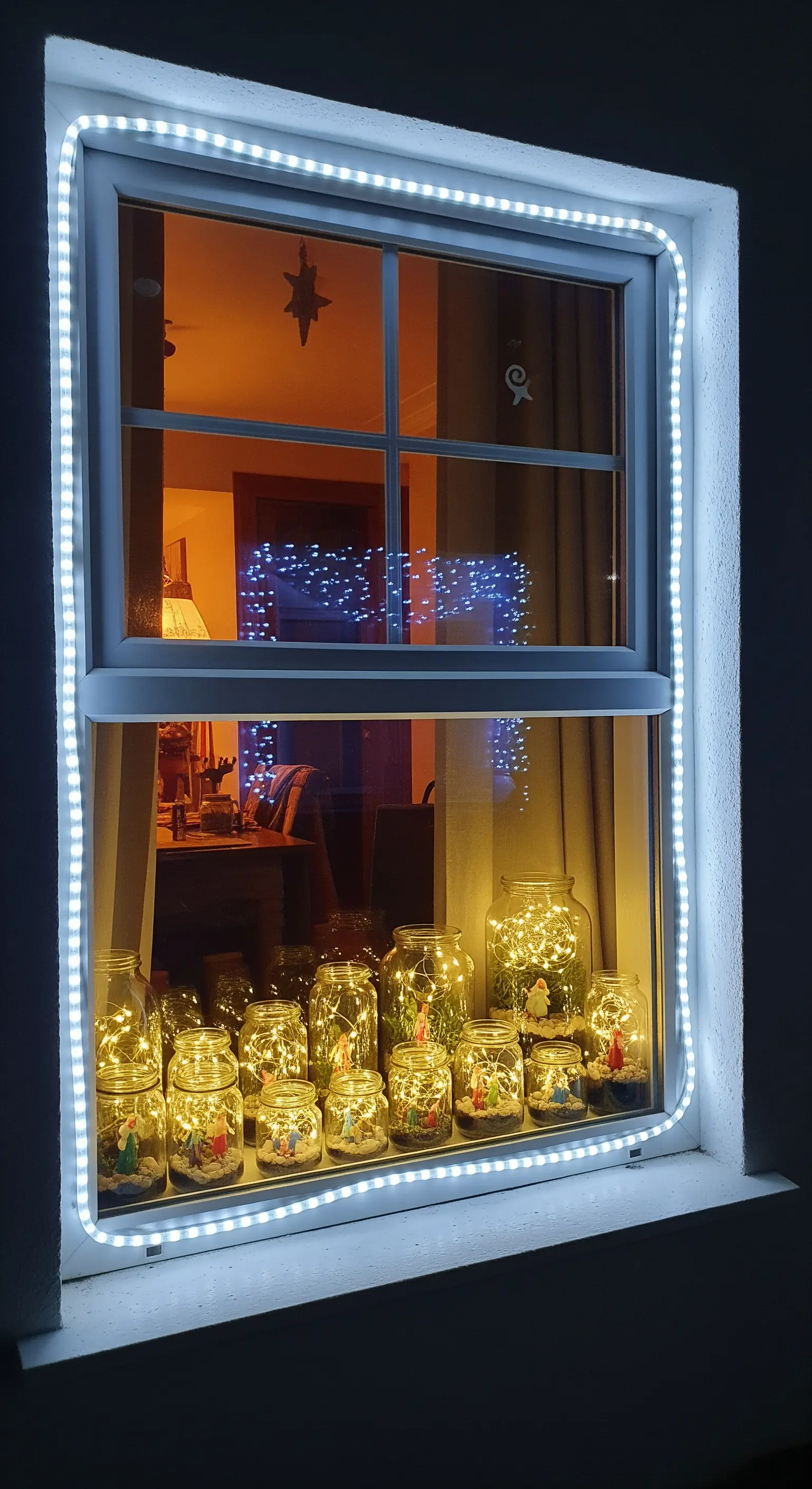 Fenster mit weißem LED-Lichtschlauch-Rahmen und innenliegenden beleuchteten Gläsern auf der Fensterbank.