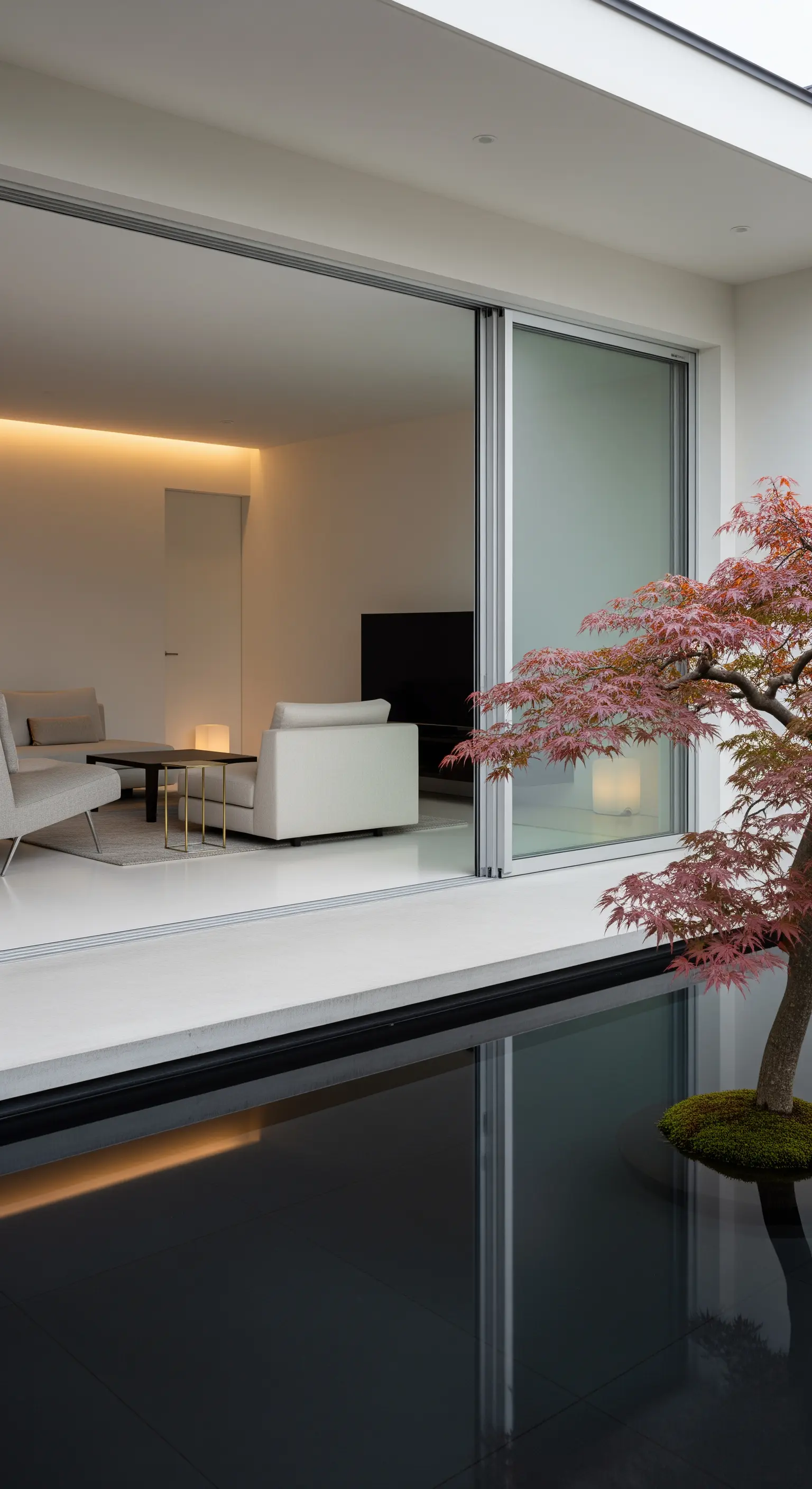 Minimalistischer Wohnraum mit Schiebetür zu einem dunklen Reflexionsbecken mit Ahornbaum.