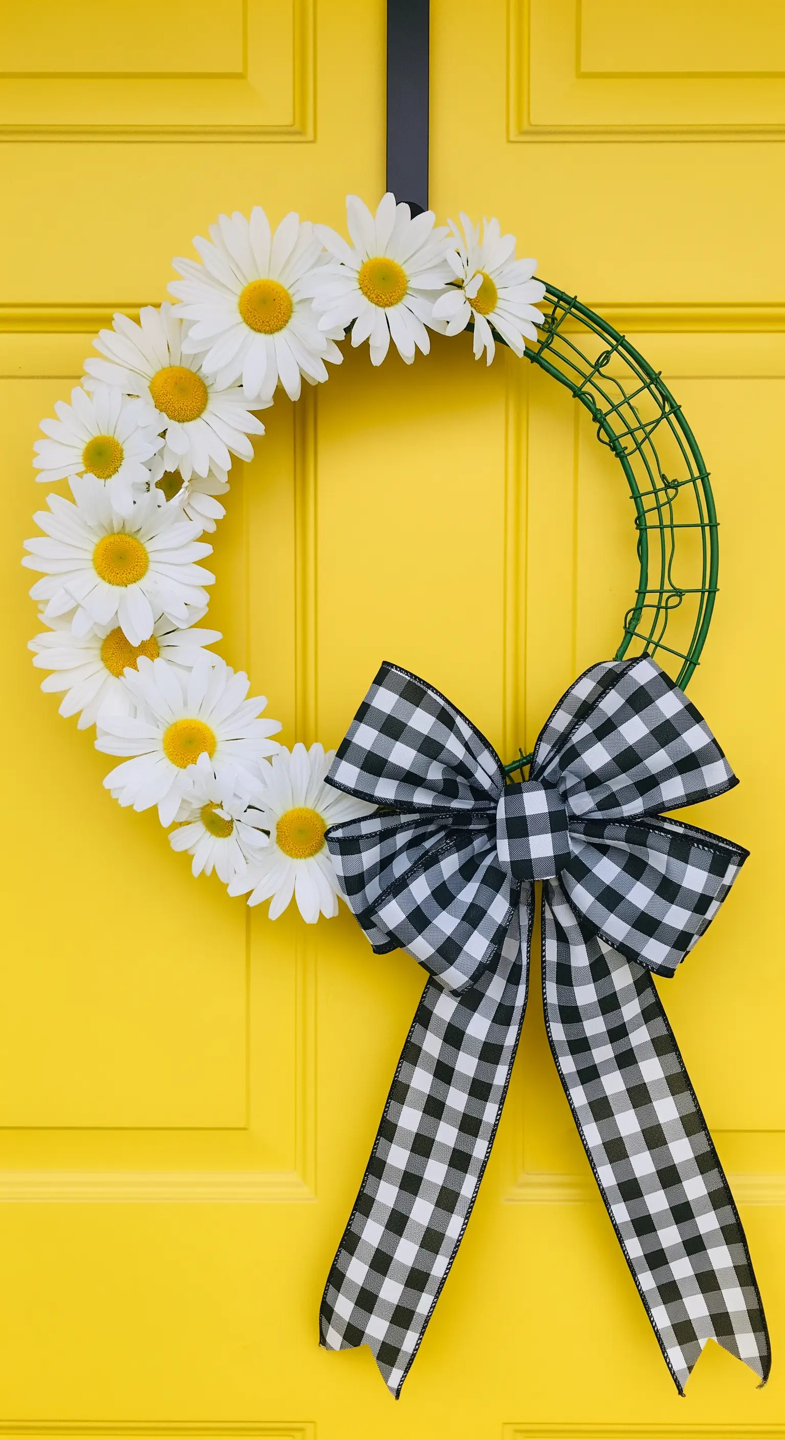Kranz aus weißen Gänseblümchen mit schwarz-weiß kariertem Band auf einer gelben Tür.