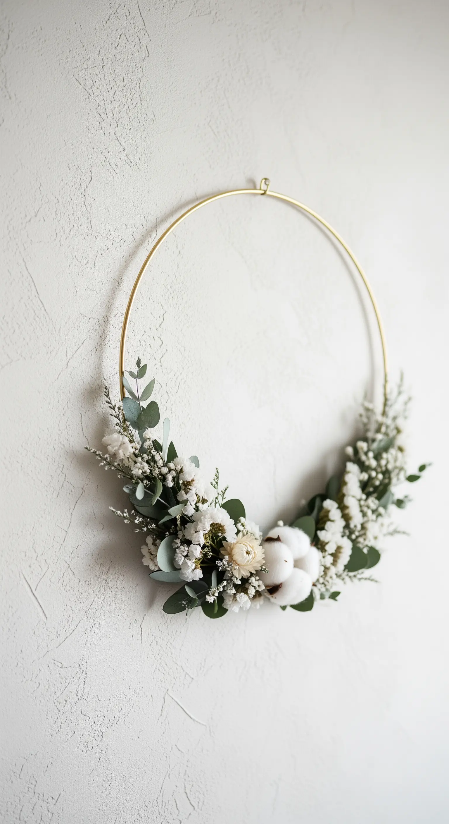 Goldener Metallring, asymmetrisch mit Eukalyptus und Baumwollblüten dekoriert, an einer weißen Wand.