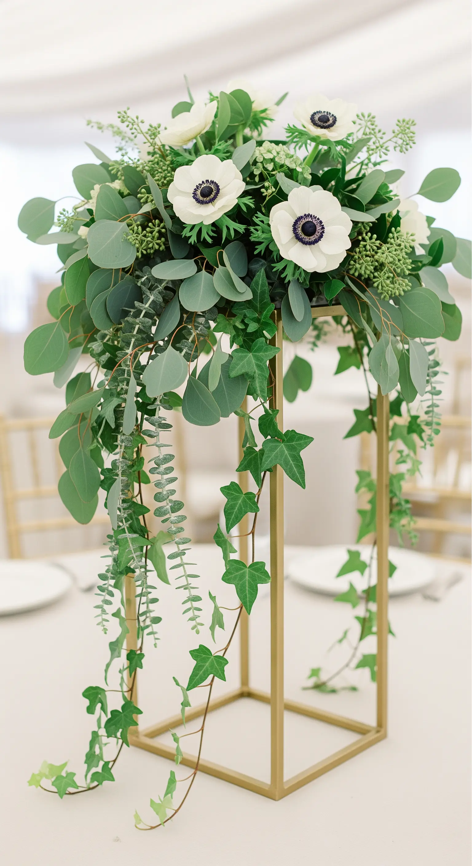 Hohe, goldene Etagere mit einem üppigen Arrangement aus Eukalyptus, Efeu und weißen Anemonen.