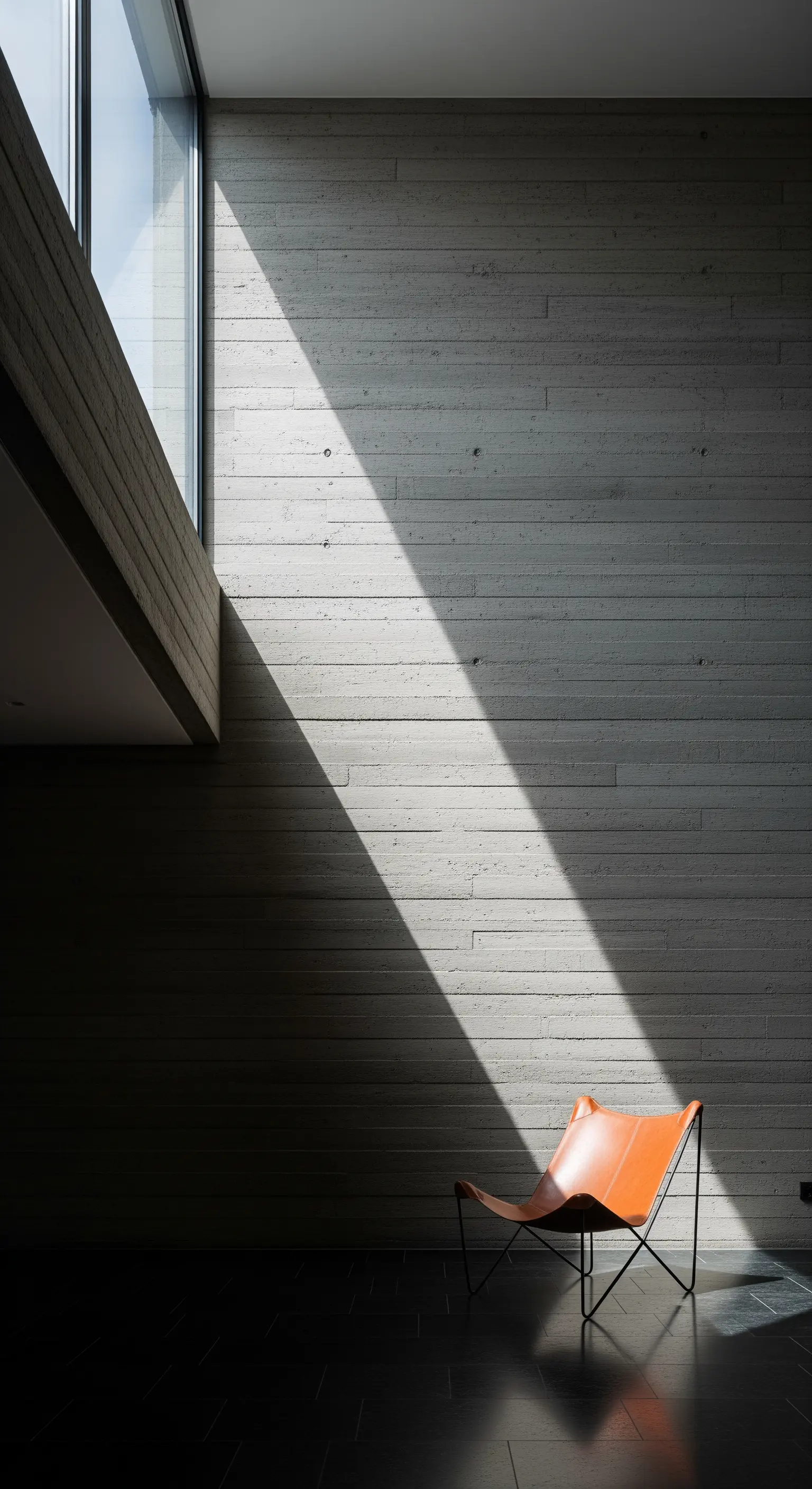 Ein Ledersessel steht im Lichtkegel, der von einem hohen Fenster auf eine Betonwand fällt.