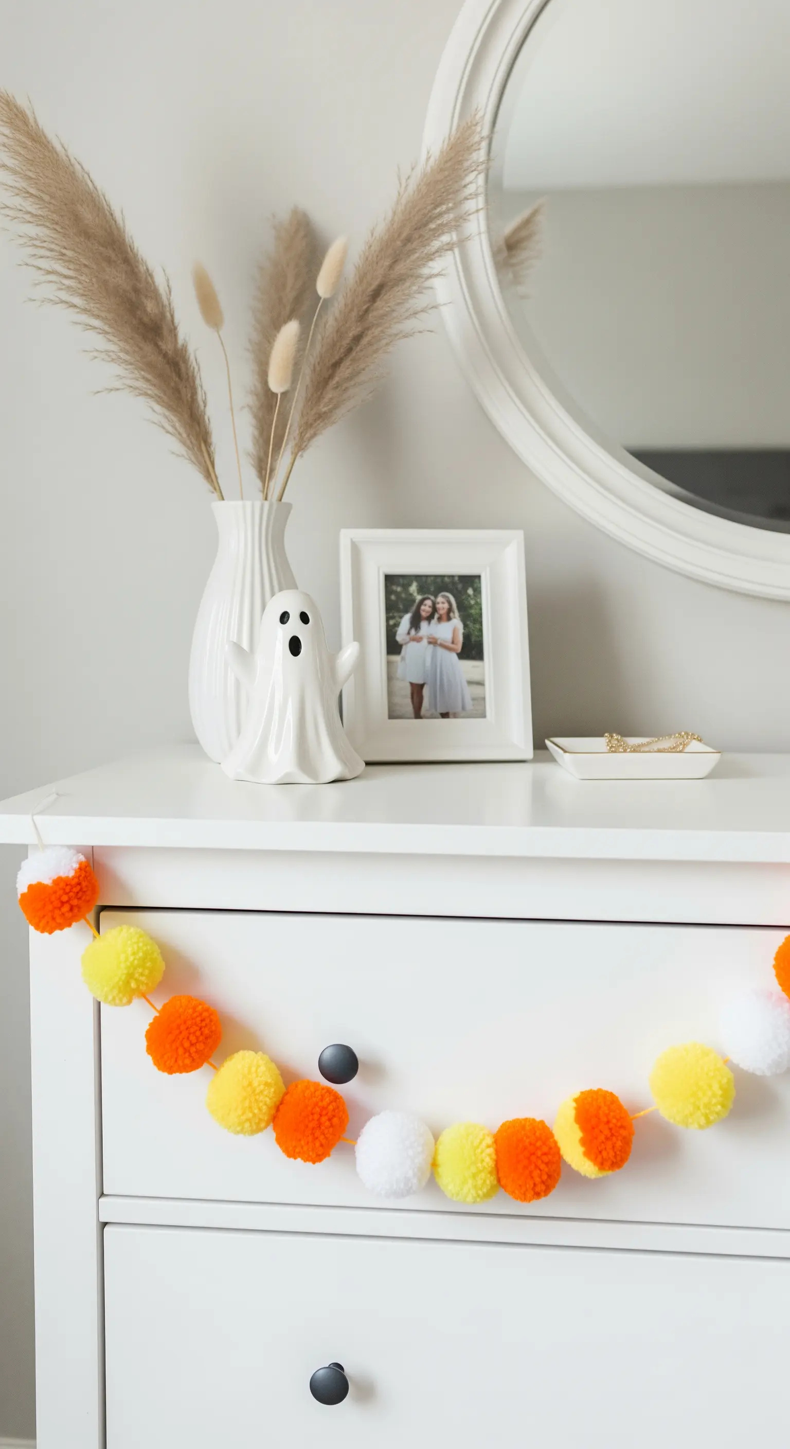 Eine weiße Kommode, dekoriert mit einer Bommelgirlande, einer Vase mit Pampasgras und einer Geisterfigur.