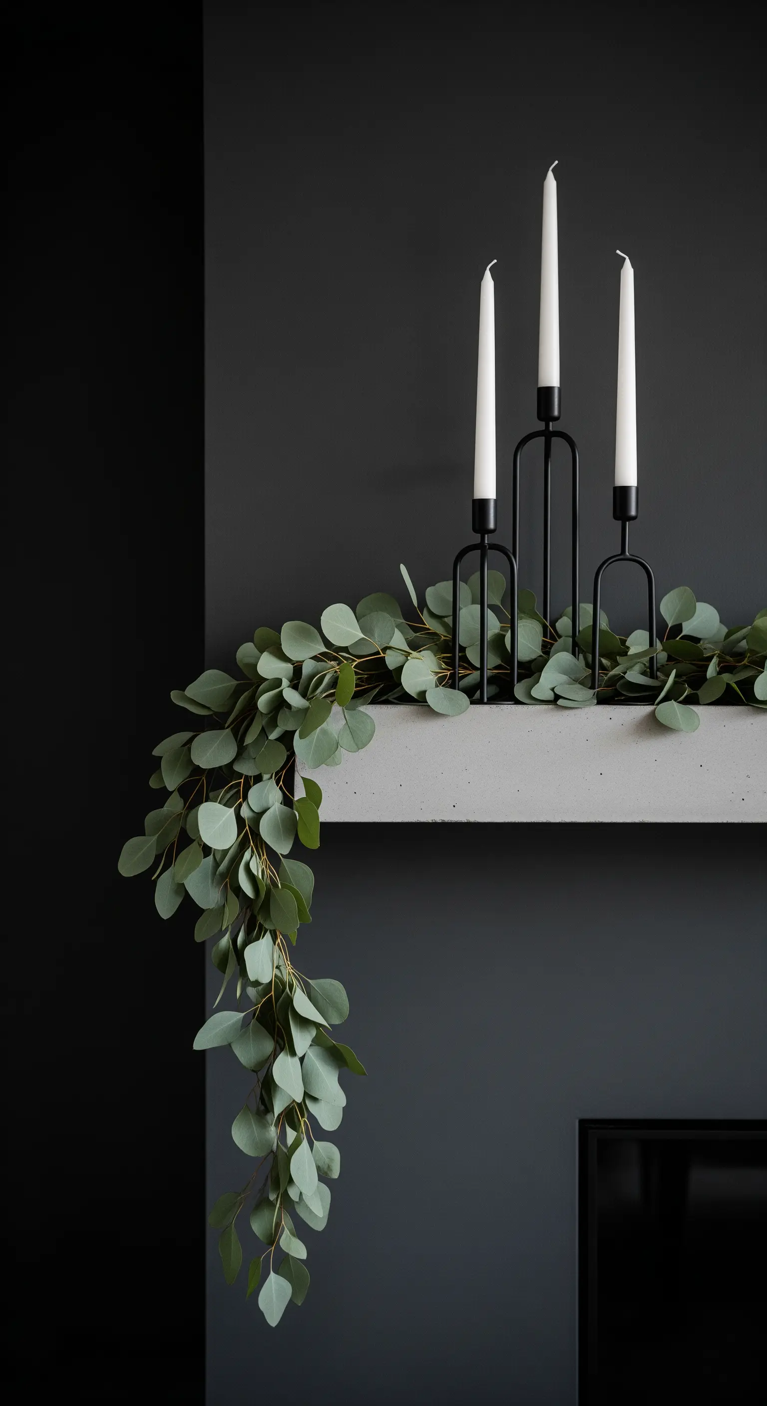 Minimalistische Deko mit einer Eukalyptus-Girlande und schwarzen Kerzenhaltern auf einem Beton-Kamin.