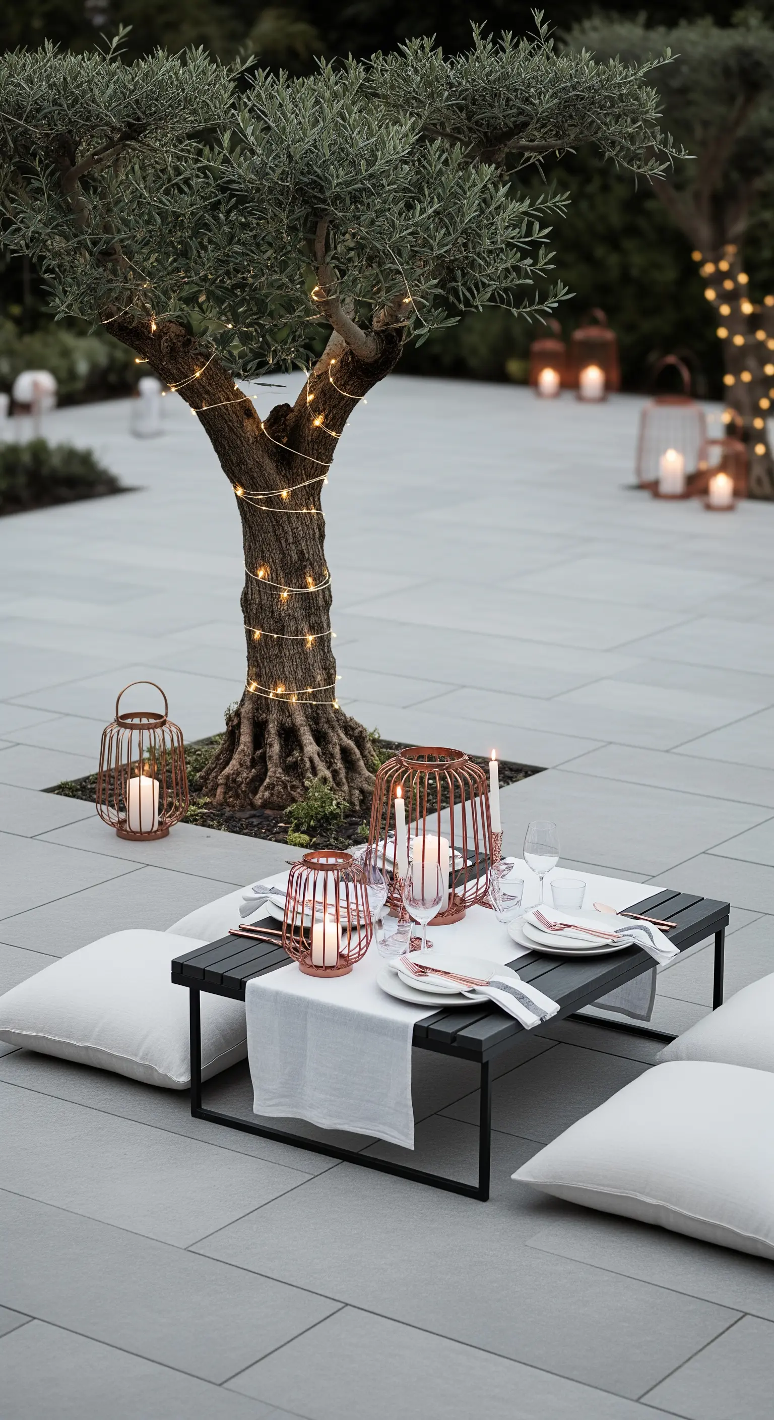 Moderne Terrasse mit Olivenbaum, dunklem Tisch, weißen Kissen und Kupfer-Laternen