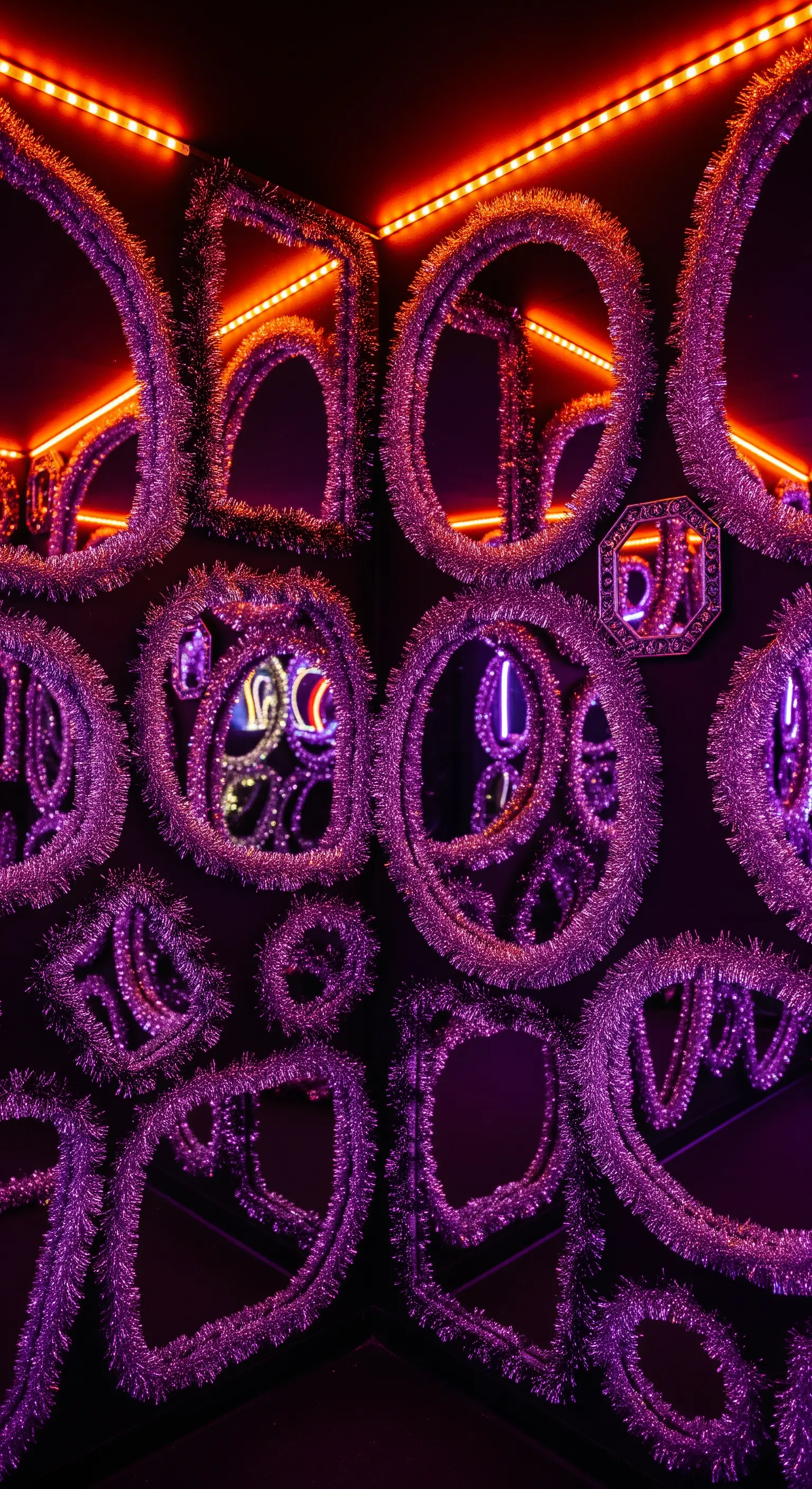 Wand voller verschiedener Spiegel mit lila Lametta-Rahmen und orangefarbener Neonbeleuchtung