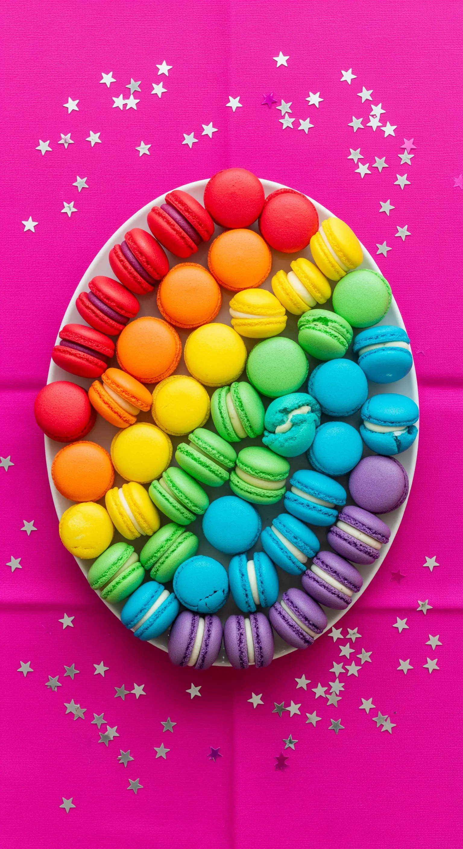Regenbogen-Macarons kreisförmig auf weißer Platte auf pinkfarbenem Tuch mit Glitzersternen.