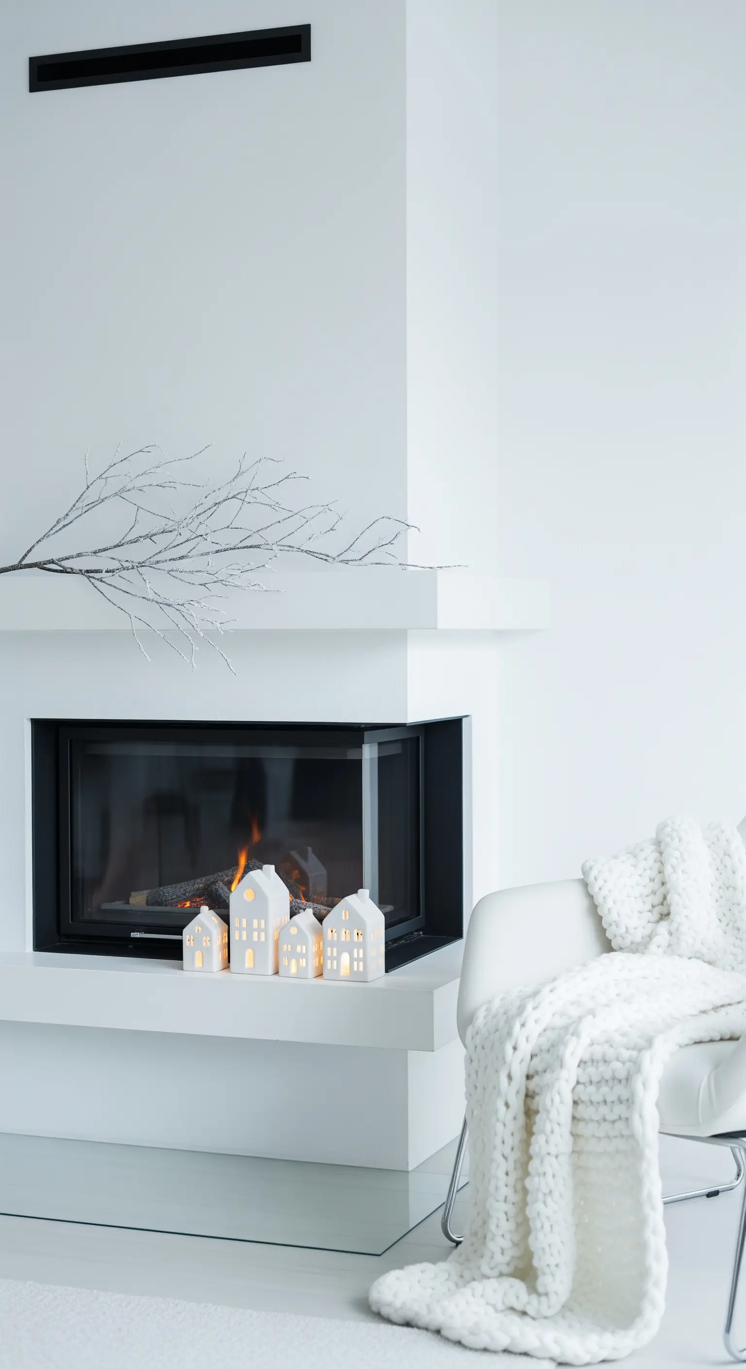 Moderner weißer Kamin mit leuchtenden Keramikhäusern und einem schneebedeckten Ast.