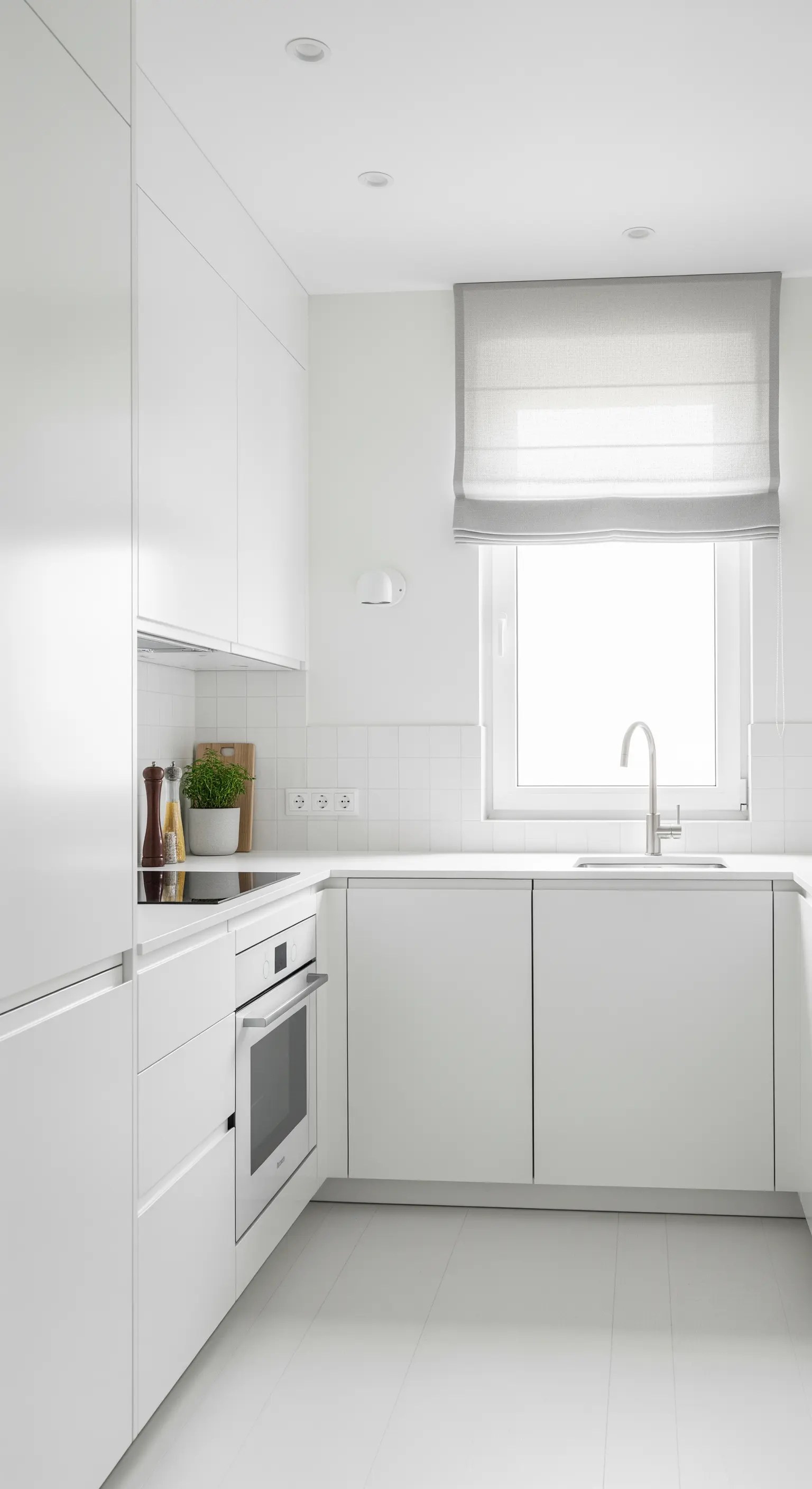 Komplett weiße, minimalistische Küche in einem schmalen Raum mit hellgrauem Raffrollo.