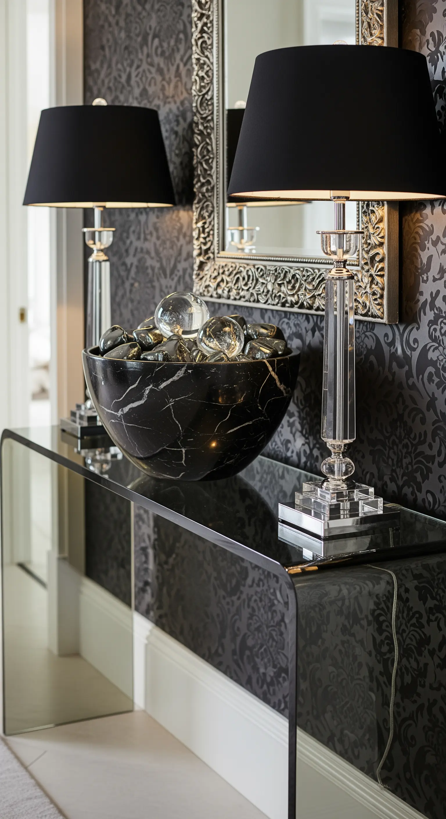 Konsole aus gebogenem Rauchglas vor einer Mustertapete, darauf eine Marmorschale und Kristalllampen.